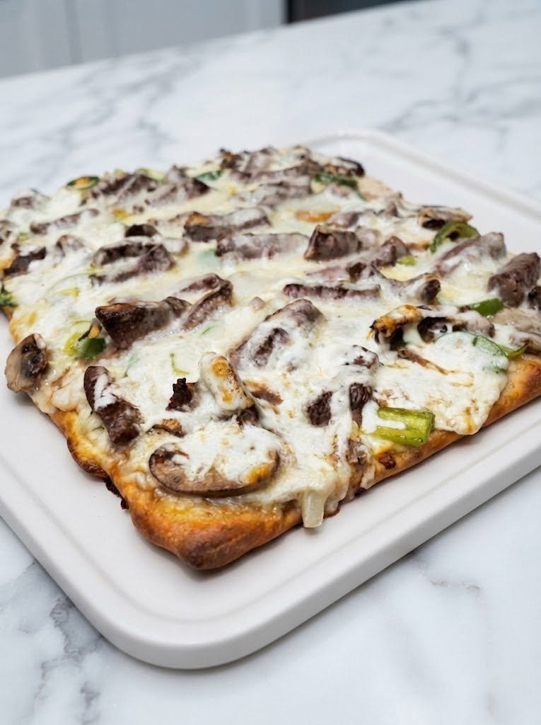 Keto Philly Cheesesteak Pizza There are some food combinations that just belong together—steak, onions, peppers, and lots of melty cheese are definitely on that list. I started thinking about how good those flavors would be piled onto a pizza, and honestly, I couldn’t stop thinking about it. So instead of reaching for a regular pizza crust, I made a Keto Philly Cheesesteak Pizza with a simple low-carb crust and loaded it with thinly sliced beef, sautéed peppers and onions, and plenty of gooey provolone and mozzarella. The first time I made it, the kitchen smelled like a cheesesteak shop. Everyone kept wandering in asking when dinner would be ready! Traditional Philly cheesesteak flavors commonly center around thin-sliced beef, onions, peppers, and melted cheese, while keto versions simply swap out the carb-heavy bread for a low-carb base. And that’s exactly what makes this pizza so satisfying. You still get the savory beef, buttery vegetables, and stretchy cheese you’re craving—just in pizza form. ❤️ Why You’ll Love This Keto Pizza Big cheesesteak flavor in every bite Low-carb and keto-friendly when using suitable ingredients Loaded with protein and cheese No traditional pizza dough required Great for weeknight dinners Easy to customize with mushrooms, jalapeños, or extra cheese Perfect when you're craving pizza but want something different 🍕 What Is Philly Cheesesteak Pizza? Philly cheesesteak pizza is basically the best of two comfort foods in one. Instead of tomato sauce and traditional pizza toppings, the crust gets a creamy garlic-cheese base, followed by thinly sliced steak, sautéed onions and bell peppers, and a generous layer of cheese. For this keto version, we're keeping the same delicious idea while using a low-carb crust. The result is cheesy, savory, hearty, and incredibly comforting. 🛒 Ingredients For the Keto Pizza Crust 2 cups shredded mozzarella cheese 2 tablespoons cream cheese 1 large egg 3/4 cup almond flour 1/2 teaspoon garlic powder 1/2 teaspoon Italian seasoning 1/4 teaspoon salt For the Cheesesteak Topping 1 pound thinly sliced ribeye or sirloin steak 1 tablespoon olive oil 1/2 medium green bell pepper, thinly sliced 1/2 medium yellow onion, thinly sliced 2 cloves garlic, minced 1 tablespoon Worcestershire sauce 1/2 teaspoon salt 1/4 teaspoon black pepper For the Creamy Pizza Base 3 tablespoons cream cheese, softened 2 tablespoons heavy cream 1/2 teaspoon garlic powder 1/4 teaspoon black pepper For the Cheese Topping 1 cup shredded mozzarella 4–5 slices provolone cheese 2 tablespoons grated Parmesan, optional 👩‍🍳 How to Make Keto Philly Cheesesteak Pizza Step 1: Prepare the oven Preheat your oven to 425°F (220°C). Line a baking sheet or pizza pan with parchment paper. If you're using a pizza stone, let it preheat in the oven. Step 2: Make the keto crust Place the mozzarella and cream cheese in a microwave-safe bowl. Heat in short 20–30 second intervals, stirring between each one, until the cheese is melted and smooth. Let it cool for a minute or two so it's easier to handle. Add the egg, almond flour, garlic powder, Italian seasoning, and salt. Mix everything together until a soft dough forms. If the mixture feels very sticky, let it sit for a couple of minutes. The almond flour will absorb some of the moisture. Step 3: Shape the crust Place the dough onto your parchment-lined baking sheet. Using lightly greased hands or another piece of parchment, press the dough into a round pizza crust about 1/4-inch thick. You can make it thicker if you prefer a softer crust or thinner if you want extra crispiness. Try to keep the thickness fairly even so it cooks evenly. Step 4: Pre-bake the crust Bake the crust for approximately 10–12 minutes, or until it looks set and lightly golden around the edges. Remove it from the oven and let it rest while you prepare the topping. 🥩 Make the Cheesesteak Filling Step 5: Cook the vegetables Heat olive oil in a large skillet over medium-high heat. Add the sliced onions and bell peppers. Cook for about 4–5 minutes, stirring occasionally, until they begin to soften. Add the minced garlic and cook for another 30 seconds. Transfer the vegetables to a plate. Step 6: Cook the steak Increase the heat slightly and add the thinly sliced steak to the same skillet. Season with salt and black pepper. Cook for approximately 3–5 minutes, stirring occasionally, until browned and cooked through. For the most tender texture, don't overcrowd the skillet. If necessary, cook the steak in batches. Add the Worcestershire sauce and stir. Return the peppers and onions to the pan and toss everything together. Cook for another minute. Remove from the heat. 🧄 Make the Creamy Pizza Sauce In a small bowl, stir together: softened cream cheese heavy cream garlic powder black pepper Mix until smooth. If the cream cheese is difficult to spread, warm the mixture for a few seconds and stir again. This creamy layer gives the pizza an extra-rich finish without needing traditional tomato sauce. 🧀 Assemble the Pizza Step 7: Add the sauce Spread the creamy garlic mixture over the pre-baked crust. Leave a small border around the edges. Step 8: Add the cheesesteak mixture Spread the cooked steak, peppers, and onions evenly over the pizza. Don't worry if it looks generously loaded. That's exactly what we want! Step 9: Add the cheese Top everything with shredded mozzarella. Arrange the provolone slices over the top. Finish with a little Parmesan if you're using it. 🔥 Step 10: Bake Return the pizza to the oven. Bake for 8–12 minutes, or until the cheese is completely melted, bubbly, and lightly golden. For an extra-browned cheesy top, switch the oven to broil for the final 1–2 minutes. Watch it closely because cheese can go from golden to burnt very quickly. Let the pizza rest for about 5 minutes before slicing. That short resting time helps the crust firm up and makes the pizza much easier to serve. ✨ My Best Tips for Perfect Cheesesteak Pizza Slice the steak very thin Thin steak cooks quickly and gives you that classic cheesesteak texture. If your steak is difficult to slice, place it in the freezer for about 20–30 minutes first. It becomes much easier to cut into thin strips. Don't overcook the beef Because the steak is sliced so thinly, it doesn't need much time. Cook it just until browned and cooked through. Don't skip the pre-bake The crust needs time to set before you add the wet toppings and cheese. This helps prevent a soggy pizza. Use provolone Mozzarella gives you that classic pizza cheese pull, but provolone brings the unmistakable cheesesteak flavor. Using both gives you the best of both worlds. Let the pizza rest I know it's tempting to cut into it immediately. Give it five minutes. Trust me—the slices will hold together much better. 🧅 Easy Variations Mushroom Philly Cheesesteak Pizza Add 1 cup thinly sliced mushrooms to the skillet with the onions and peppers. Cook until the mushrooms are browned and tender. Spicy Philly Pizza Add sliced jalapeños or a pinch of red pepper flakes to the steak mixture. You can also drizzle a little hot sauce over the finished pizza. Extra Cheesy Add another layer of provolone underneath the mozzarella. If you love cheese, there's really no reason to hold back. Garlic Butter Crust Brush the edges of the baked crust with melted butter mixed with garlic powder and parsley before serving. Ground Beef Version If thin-sliced steak isn't available, you can use ground beef. Ground beef is a convenient alternative for cheesesteak-style recipes and cooks quickly; it also works particularly well with peppers, onions, and melted cheese. 🥗 What to Serve With Keto Philly Cheesesteak Pizza This pizza is filling enough to serve on its own, but I like pairing it with something fresh and crunchy. Try: Simple green salad Caesar salad Roasted broccoli Garlic butter green beans Cucumber salad Avocado salad Air-fried zucchini Crispy Parmesan zucchini chips For a casual weekend dinner, a big salad and this pizza make a pretty perfect combination. 🧊 Make-Ahead Instructions You can prepare several components ahead of time. Cook the steak, peppers, and onions up to 2 days ahead and store them covered in the refrigerator. The cream sauce can also be prepared ahead and refrigerated. When you're ready to make dinner, prepare the crust, assemble the pizza, and bake. This makes the recipe especially convenient for busy weeknights. 🥡 How to Store Leftovers Let the pizza cool before transferring it to an airtight container. Store in the refrigerator for up to 3–4 days. For the best texture, reheat slices in: Air fryer Toaster oven Conventional oven Heat at about 350°F until warmed through and the cheese is bubbly again. The microwave works when you're in a hurry, but the crust won't be as crisp. ❄️ Can You Freeze Keto Philly Cheesesteak Pizza? Yes. Let the baked pizza cool completely. Slice it into individual portions and wrap each slice well before placing it in a freezer-safe container or bag. Freeze for up to 2 months for the best quality. Reheat from frozen in an oven or air fryer until hot and crisp. ❓ Frequently Asked Questions Is Philly cheesesteak pizza keto? This version is designed to be keto-friendly by replacing traditional wheat pizza dough with a low-carb almond-flour and cheese crust. Be sure to check the labels on your cheese, sauces, and other packaged ingredients if you track carbohydrates closely. What steak is best for Philly cheesesteak pizza? Ribeye is my first choice because it's flavorful and tender. Sirloin is another good option if you prefer something a little leaner. Can I use leftover steak? Absolutely. Thinly slice cooked steak and warm it briefly with the peppers, onions, and Worcestershire sauce before assembling the pizza. Can I use ground beef? Yes. Brown the ground beef first, then add the onions, peppers, garlic, and seasonings. Ground beef is also a popular shortcut for keto cheesesteak-style meals. Can I use only mozzarella? Yes, but provolone adds a more classic cheesesteak character. A combination of mozzarella and provolone gives you excellent flavor and melt. Why is my keto crust soft? Keto cheese-based crusts can be softer than traditional flour crusts. Bake the crust until set before adding toppings, avoid piling on excessive sauce, and allow the finished pizza to rest before slicing. Can I make the crust thinner? Definitely. A thinner crust will become crispier and more pizza-like. Just keep an eye on it during the pre-bake because a thin crust can brown quickly. Can I make this without bell peppers? Of course. If you're going for a more minimalist cheesesteak flavor, simply use steak, onions, and cheese. 📝 Recipe at a Glance Prep Time: 20 minutes Cook Time: 25 minutes Total Time: About 45 minutes Servings: 4–6 Course: Dinner Cuisine: American-inspired / Keto Ingredients Crust 2 cups mozzarella 2 tbsp cream cheese 1 egg 3/4 cup almond flour 1/2 tsp garlic powder 1/2 tsp Italian seasoning 1/4 tsp salt Topping 1 lb thinly sliced ribeye or sirloin 1 tbsp olive oil 1/2 green bell pepper 1/2 yellow onion 2 garlic cloves 1 tbsp Worcestershire sauce 1/2 tsp salt 1/4 tsp black pepper Creamy Base 3 tbsp cream cheese 2 tbsp heavy cream 1/2 tsp garlic powder 1/4 tsp black pepper Cheese 1 cup mozzarella 4–5 slices provolone 2 tbsp Parmesan, optional Instructions Preheat oven to 425°F (220°C). Melt mozzarella and cream cheese for the crust. Mix with egg, almond flour, garlic powder, Italian seasoning, and salt. Shape into a 1/4-inch-thick pizza crust. Pre-bake for 10–12 minutes. Sauté peppers, onions, and garlic. Cook thinly sliced steak with salt, pepper, and Worcestershire sauce. Return vegetables to the skillet and combine. Mix the creamy garlic base. Spread sauce over the crust. Add steak, peppers, and onions. Top with mozzarella and provolone. Bake 8–12 minutes, until bubbly and golden. Rest for 5 minutes, slice, and serve. ❤️ Final Thoughts This Keto Philly Cheesesteak Pizza is exactly the kind of dinner I love when I can't decide whether I want pizza or a cheesesteak. Thankfully, you don't have to choose. You get tender steak, sweet sautéed onions, colorful peppers, creamy garlic sauce, and that gorgeous blanket of melted provolone and mozzarella—all sitting on a crispy, cheesy low-carb crust. It's hearty enough for dinner, fun enough for a weekend, and satisfying enough that nobody has to feel like they're eating “diet food.” Sometimes keto cooking is all about finding clever ways to keep the foods you love. And this one is definitely a keeper.